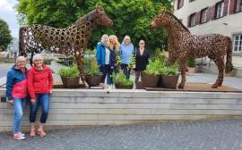 Zwei überlebensgroße Pferde aus Hufeisen stehen im Hintergrund. Davor die Frauen der Gruppe und viele Kräu