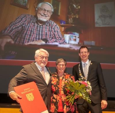 Drei Personen lächeln in die Kamera: eine Mann mit Urkunde in der rechten Hand, eine Frau mit Blumenstrauß