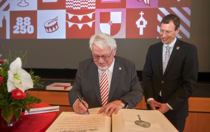 Ein Mann schreibt in ein dickes Buch, ein weiterer Mann schaut ihm dabei zu