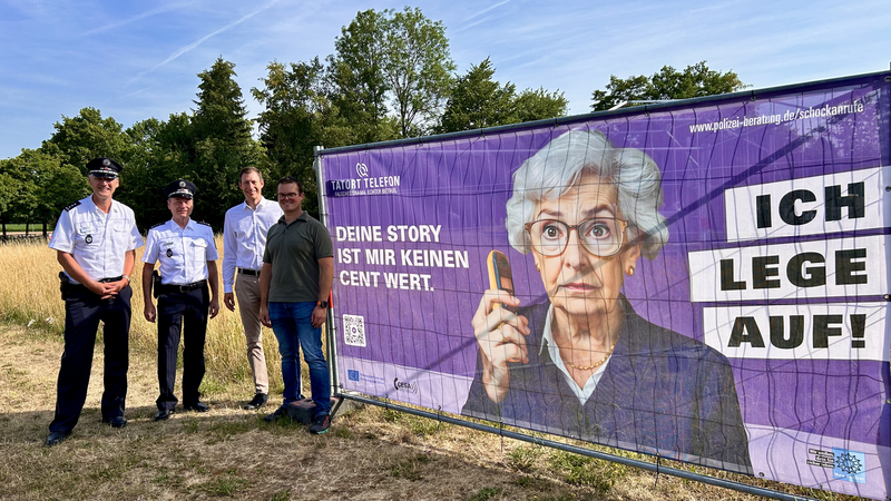 Vier Männer, davon zwei Polizisten, stehen neben einem großen lilafarbenen Plakat, auf dem eine alte Dame mit erschrockenem Gesicht ein Telefon in der Hand hält