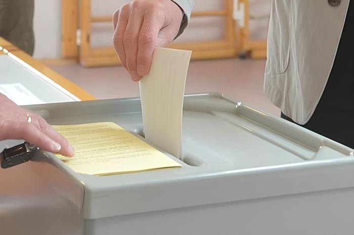 Eine Hand hält einen Stimmzettel und steckt diesen in den Schlitz der grauen Wahlurne
