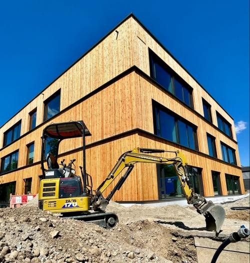 Ein mit warmem Holz verkleidetes, dreistöckiges Gebäude mit großen Fenstern, davor ein gelber Bagger