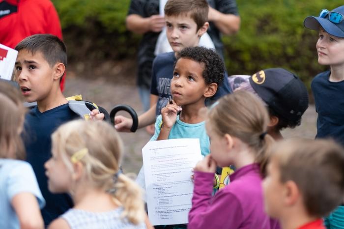 Kinder mit Zetteln in der Hand hören einem Stadtführer zu.