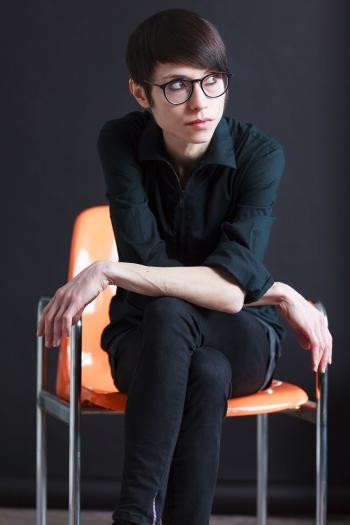 Eine Frau, ganz in Schwarz gekleidet, mit dunkler Brille und dunklem Kurzhaarschnitt, sitzt auf einem orangefarbenen Stuhl