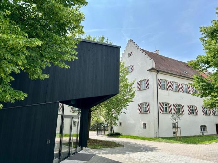 Das Gebäude links ist aus Holz und außen schwarz. Rechts ein historisches Haus mit rot-weißen Fensterläden