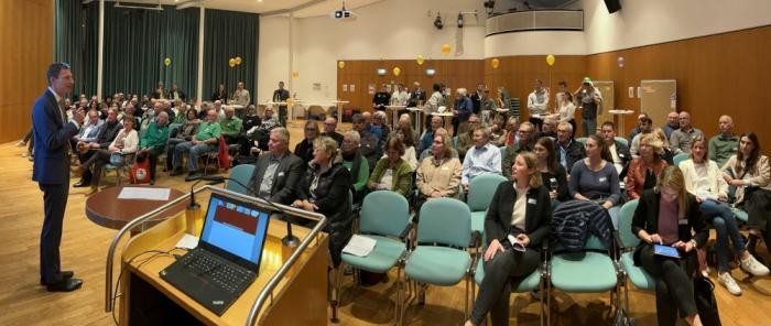 Der Oberbürgermeister steht links am Bildrand und spricht zu ca. 80 Männer und Frauen, die auf Stühlen sitzen