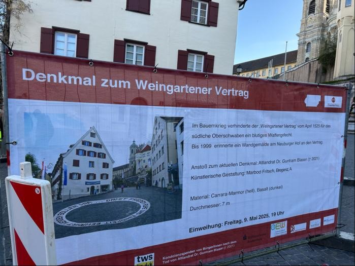 Ein Plakat in Weiß und Rot enthält Informationen, was an der Baustelle entstehet, nämlich ein kreisrundes Denkmal in den Pflastersteinen