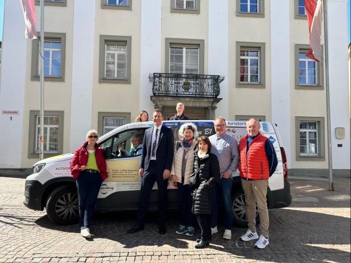 Gruppenbild vor dem Fahrzeug mit Fahrern und Fahrerinnen sowie Oberbürgermeister Clemens Moll. Im Hintergrund das Rathaus