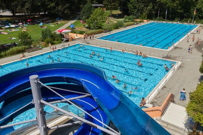 Das Freibad aus der Vogelperspektive: unten im Bild sind Teile der blauen Riesenrutsche zu sehen, hinten das Nichtschwimmer- und das Schwimmerbecken mit einigen Gästen im Wasser