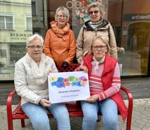 Vier Frauen hinter und auf einer roten Sitzbank. Sie halten ein Plakat mit dem Hinweis "Hinsetzen. Schwätzen" in den Händen