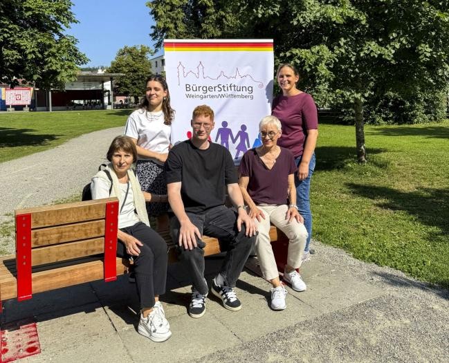 Fünf Persoen sitzen auf oder stehen neben einer Holzbank im Park