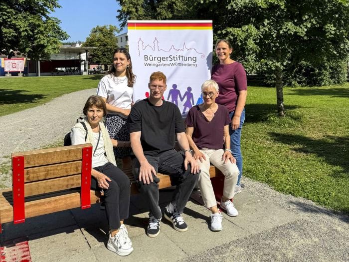 Fünf Personen stehen neben einer Bank oder sitzen auf ihr. Drumherum Wiese und Bäume.