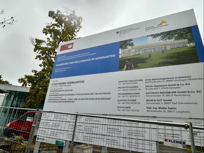 Ein großes Schild mit vielen Daten und Firmennamen steht vor einer Baustelle