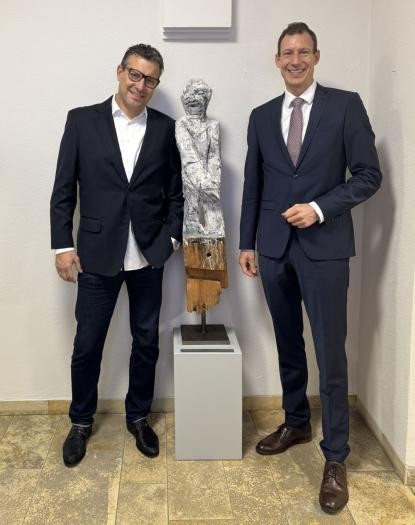 Zwei Männer stehen vor einer Wand, zwischen ihnen die fast mannshohe Holzskulptur