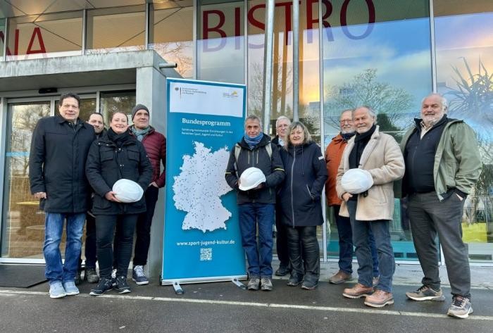 Gruppenbild mit zehn Personen, von denen manche einen weißen Bauhelm in der Hand halten. Im Hintergrund die Glasfassade eines Gebäudes
