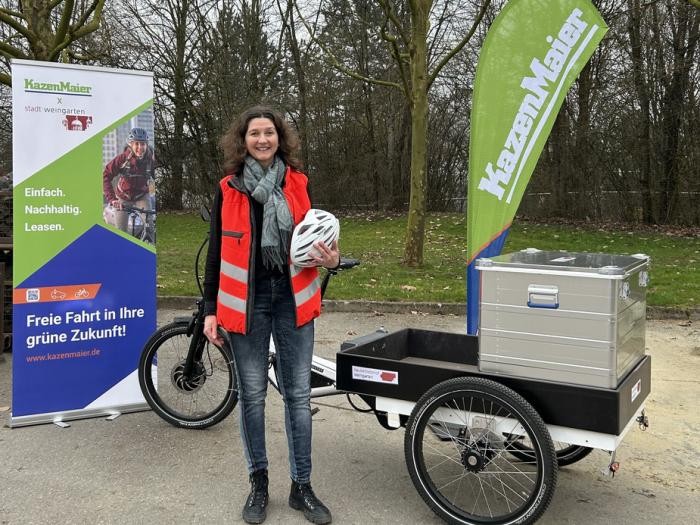 Eien Frau mit Fahrradhelm steht vor einem Fahrrad mit drei Rädern und Anhänger