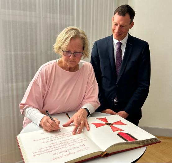 Eine Frau schreibt etwas in ein großes Buch, ein Mann sieht ihr dabei zu.