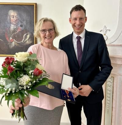 Eine Frau mit Blumen und Urkunde lächelt in die Kamera, neben ihr ein Mann in Anzug und Krawatte. Beide in einem festlichen Saal