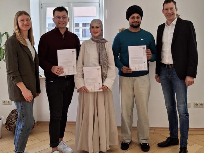 Fünf Menschen stehen in einer Reihe und lächeln in die Kamera. Drei von ihnen halten ein Papier in der Hand. 