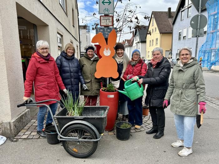 Sieben Frauen mit Gießkannen und Handschuhen vor einem roten Kräuterkübel