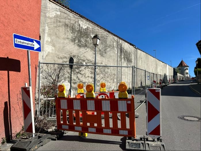 Eine lange helle Mauer mit Rissen und Flecken entlang einer Straße, davor eine Baustellen-Absperrung