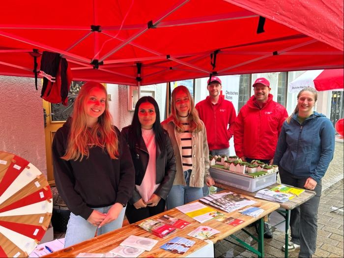 Vier Frauen und zwei Männer stehen in einer Reihe unter einem großen roten Schirm. Vor ihnen auf dem Tisch liegen Hefte und Handzettel.