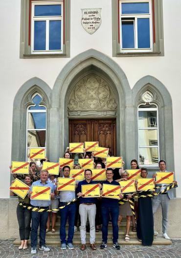 Eine große Gruppe Frauen und Männer hält vor einem Gebäude gelbe Schilder hoch, auf denen Wörter wie "Schwimmbad" oder "Parkpflege" rot durchgestrichen sind.