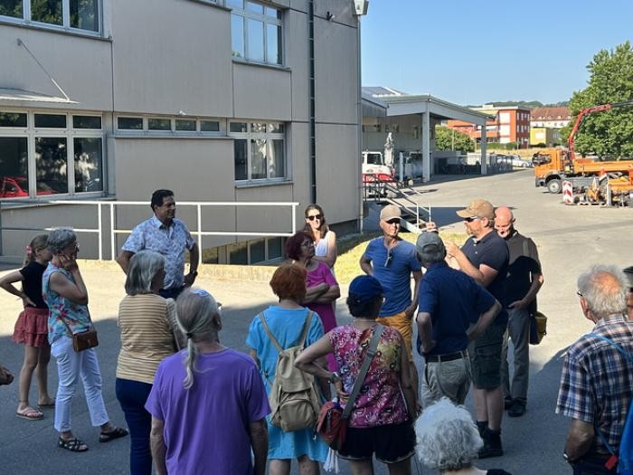 Eine Gruppe Menschen versammelt sich vor einem Gebäude um einen Mann, der gestikulierend zu ihnen spricht  