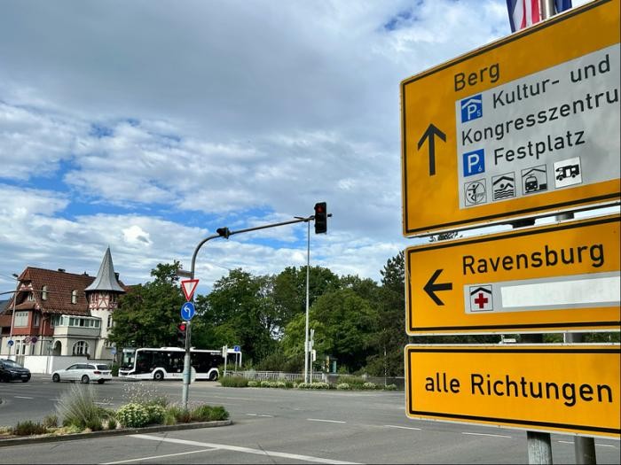 Ein gelbes Verkehrsschild mit der Pfeilrichtung nach "Ravensburg" und eine vierspurige große Kreuzung, auf der ein Linienbus zu sehen ist
