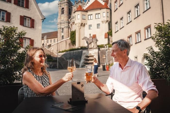 Ein Mann und eine Frau sitzen in einem Cafe auf einem Platz und prosten sich lachend mit einem Glas zu