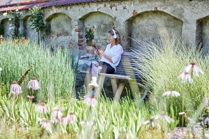 Eine Frau sitzt mit weißen Kopfhörern auf den Ohren auf einer Gartenbank mitten im Grünen
