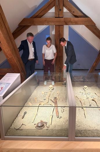 Drei Personen stehen in einem Museum vor einem Glaskasten, in dem ein Skelett liegt.
