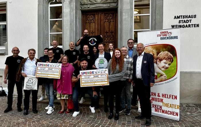 Gruppenbild mit vielen Männern und Frauen vor einem Haus. Mehrere Personen halten ein Schild in der Hand, auf dem ein Geldbetrag steht.