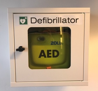 Das Gerät mit der Aufschrift "AED" befindet sich in einem Schrank mit Fenster. Oben am Fenster die Überschrift "Defibrillator"