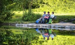 Drei Wanderer mit Rucksäcken sitzen auf einer Bank und blicken auf den Kreuzbergweiher